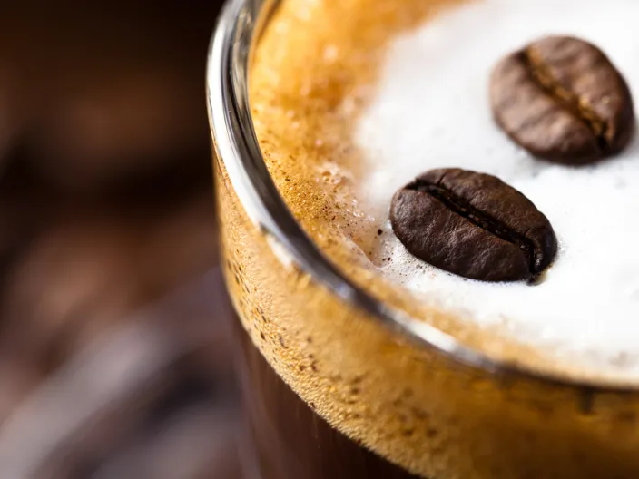 Närbild på kaffe upphällt i ett glas med två kaffebönor slutandes i det vita skummet